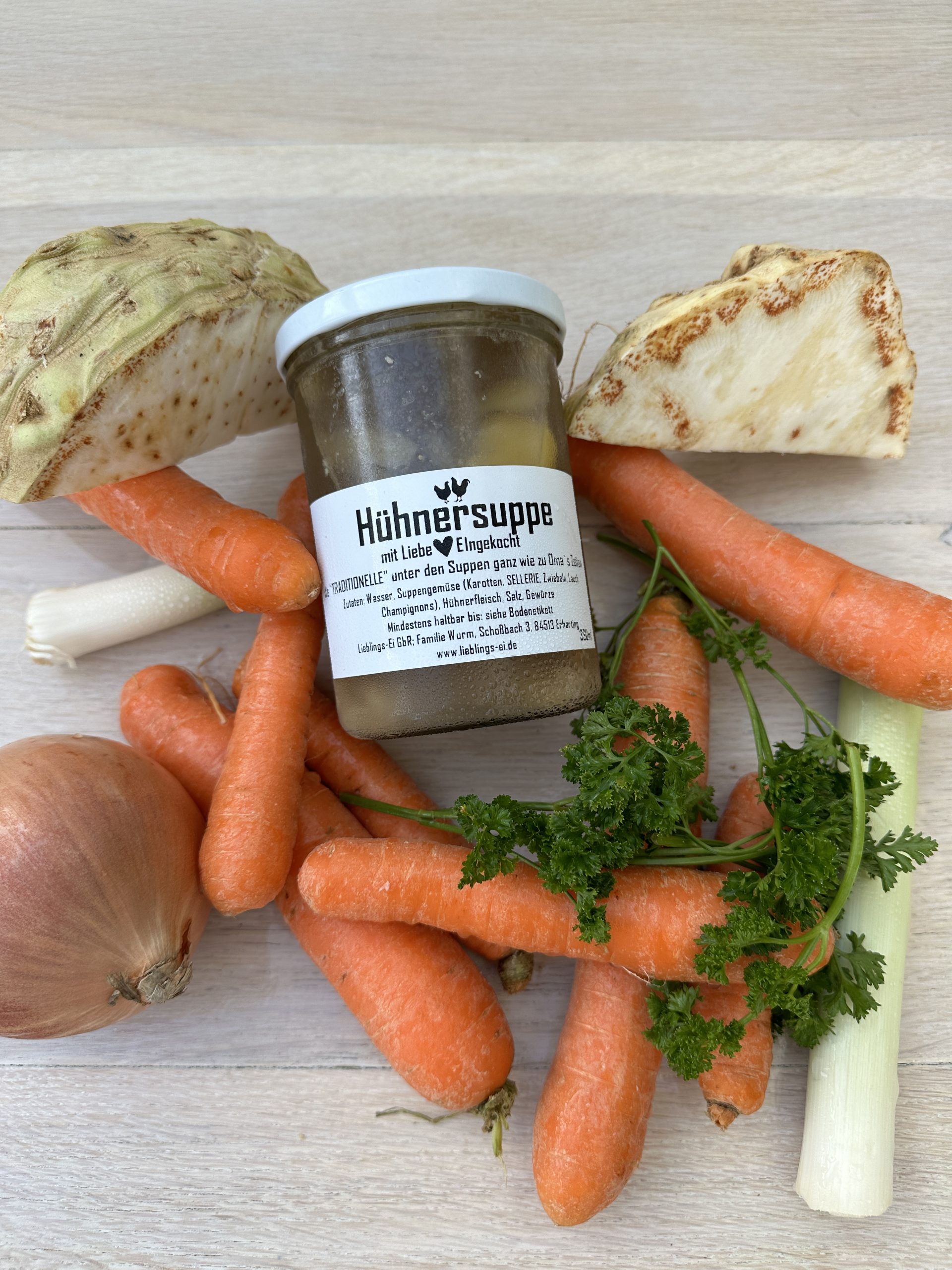 Ein Einweck-glas mit Hühnersuppe liegt zwischen frischen Karotten, Petersilie, Zwiebeln und Sellerie auf einem Holztisch